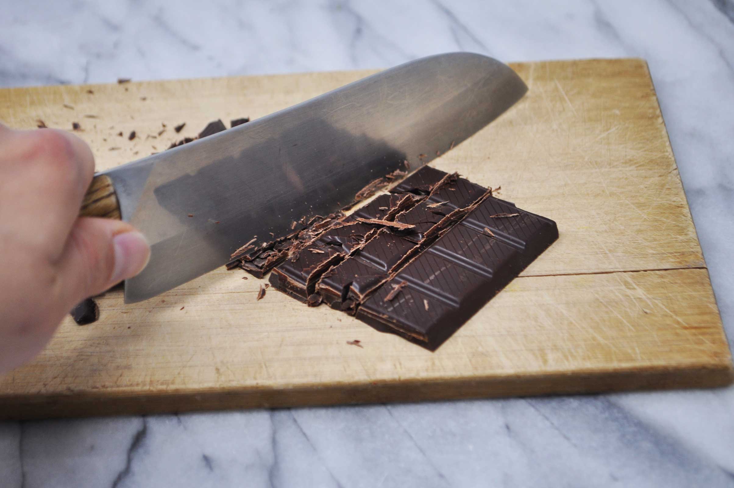 Image of a hand holding a chef's knife, cutting a bar of dark chocolate into small pieces on a cutting board